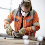 Mann in orangener Arbeitskleidung mit Baukopfhörer, Maske und Schutzbrille schleift Metallstück.