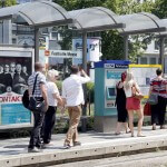 Personen laufen an der Haltestelle "Festhalle/Messe" bei sonnigem Wetter entlang