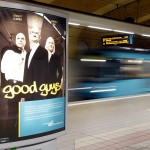 "Good Guys" Plakat an einer unterirdischen Haltestelle. Im Hintergrund eine Frau vor einer vorbeifahrenden U-Bahn