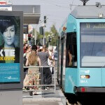 Plakat "Die Akte Ivankovic" vor einer Straßenbahnhaltestelle an der Personen in die Straßenbahn 11 einsteigen wollen.