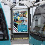 Plakat "Sisters on Rails" am Haltestellen-Häuschen zwischen zwei Wagons zu sehen