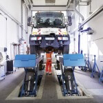 Mitarbeiter in orangener Arbeitskleidung unter VGF Fahrzeug, das auf Wagenhebern steht, in der Werkstatt