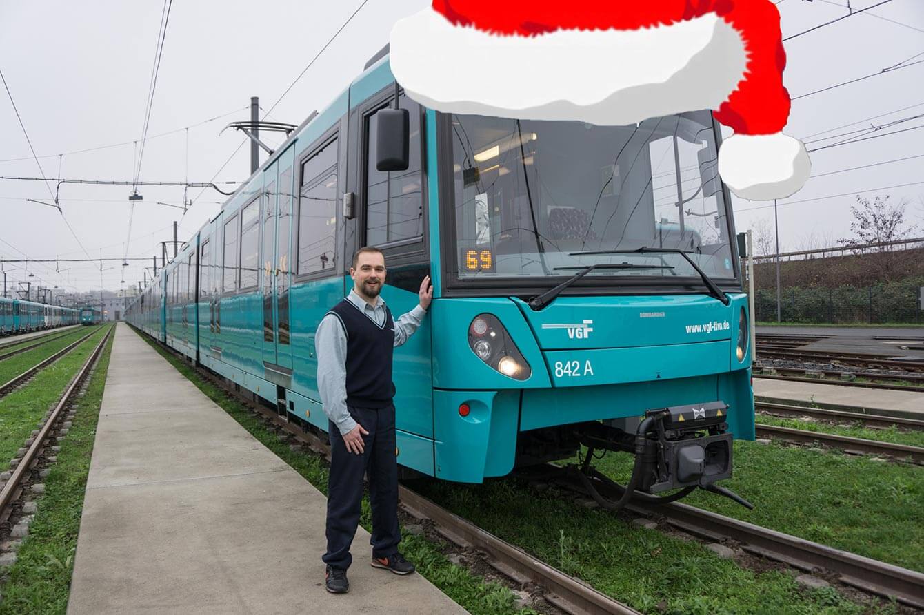 Mitarbeiter der VGF neben einer VGF Bahn auf die eine Weihnachtsmütze editiert wurde.
