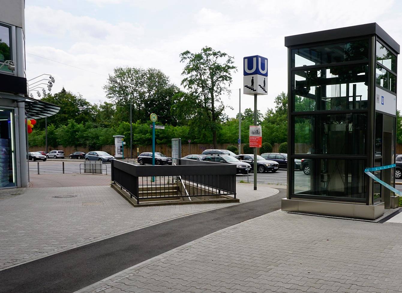 Verglaste Aufzugsanlage Miquel-/Adickesallee von außen am Eröffnungstag. Daneben die Treppe zur unterirdischen Haltestelle, ein blaues Schild mit einem "U" darauf und darunter ein weißes mit einer Person die die Treppe herunter läuft.