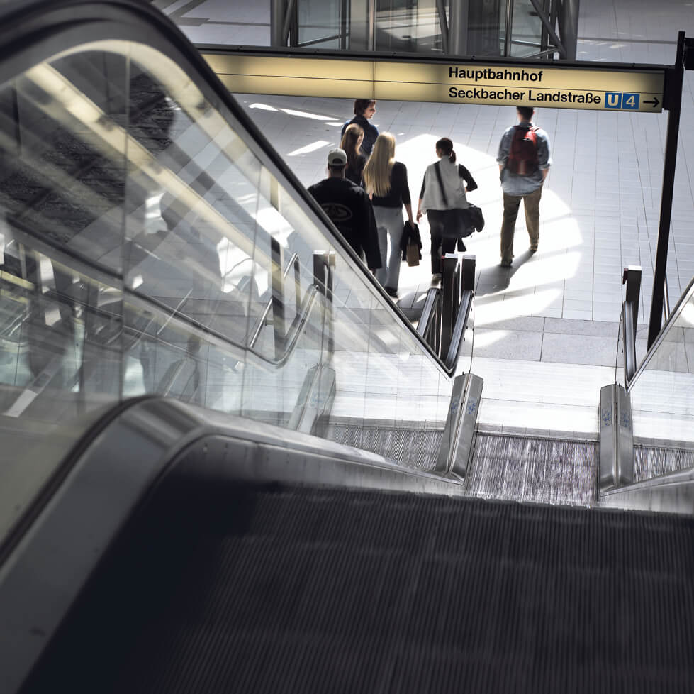 Rolltreppen von oben nach unten fotografiert. Unten laufen sechs Personen die unterirdische Haltestellen-Plattform
