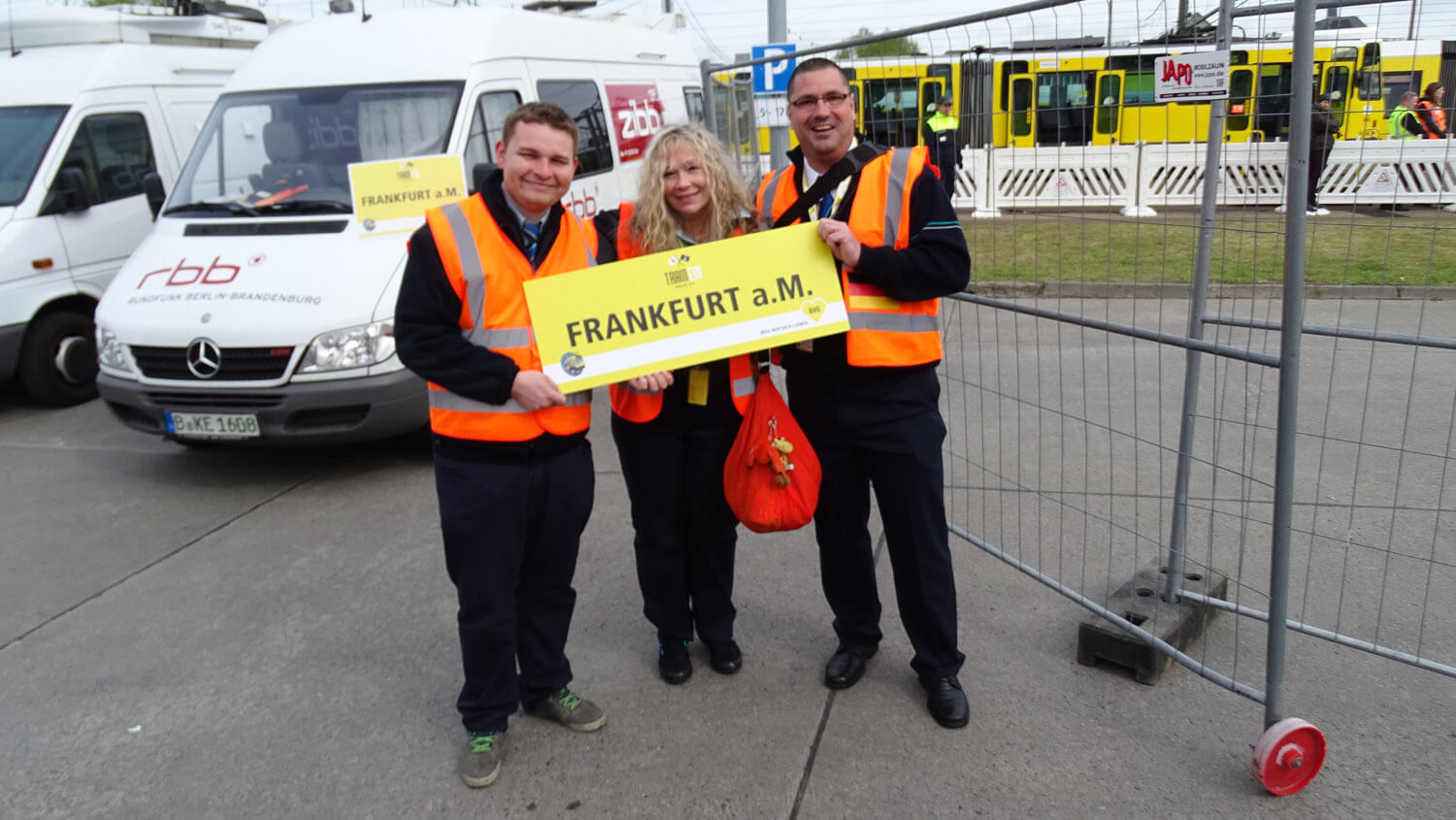 Drei Mitarbeiter der VGF tragen orangene Warnwesten und halten ein gelbes Schild "Frankfurt a.M."