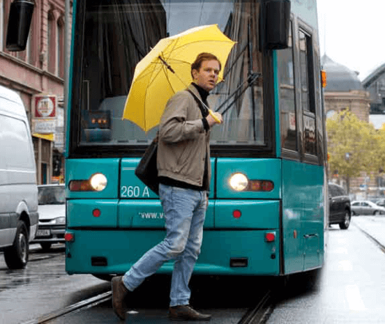 Mann mit gelben Regenschirm läuft vor einer Bahn über die Straße