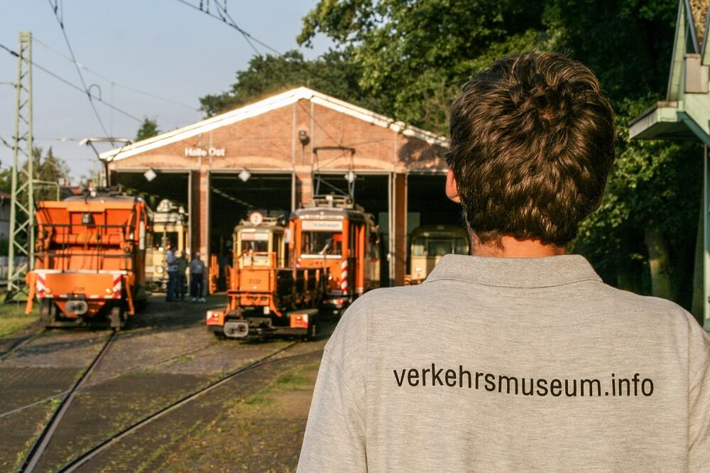 Person von hinten mit grauem Shirt mit Aufschrift auf dem Rücken "verkehrsmuseum.info" vor der Halle Ost mit Schienenbaustellenfahrzeugen.