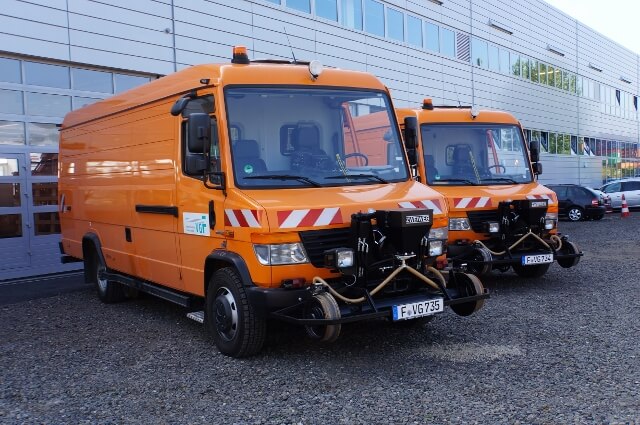 Zwei orangene Zweiwegefahrzeuge vor einem Gebäude die auf Straße und Schienen fahren können.
