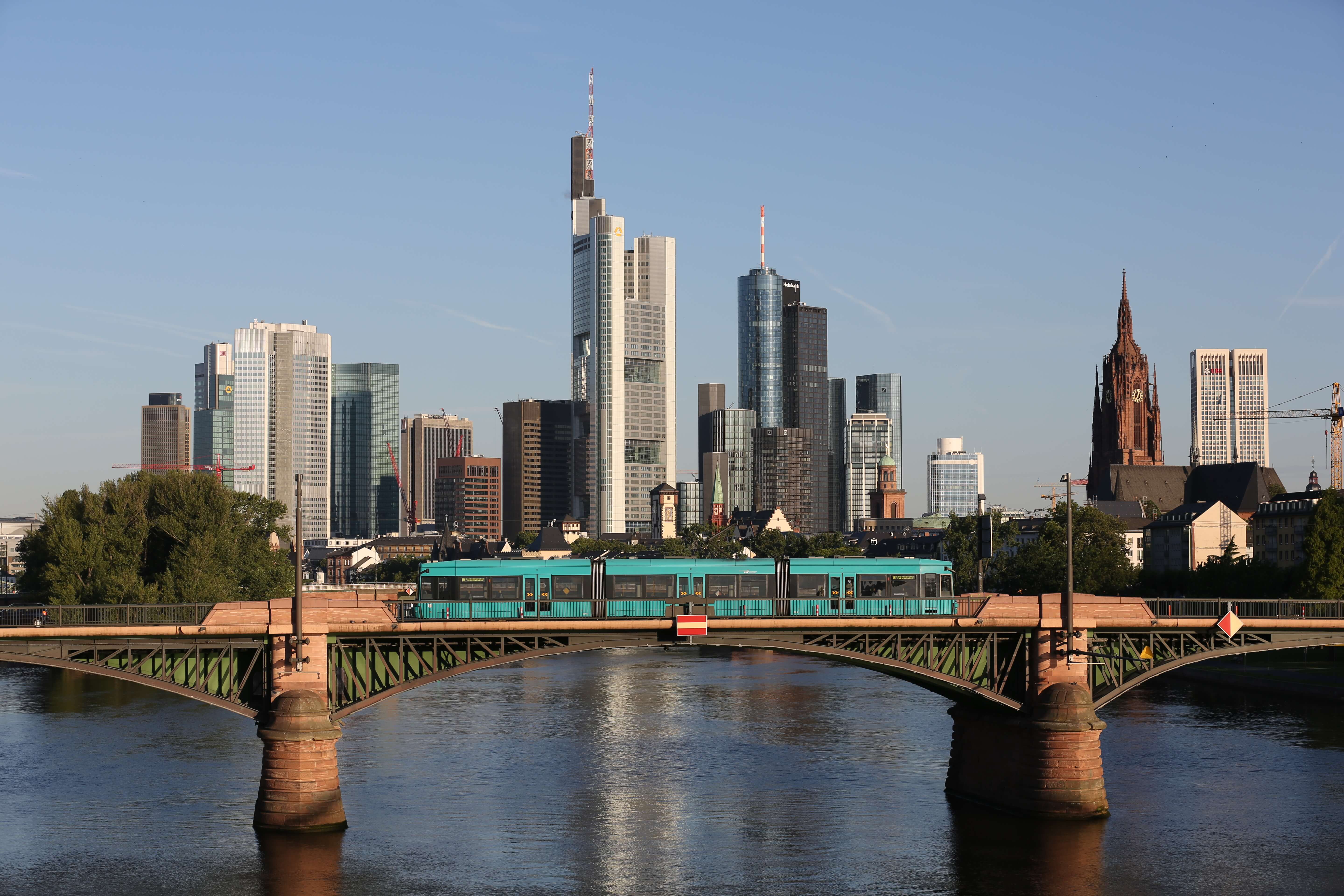 Skyline Frankfurt mit dem Main davor und einer Bahnbrücke auf der eine VGF-bahn fährt.