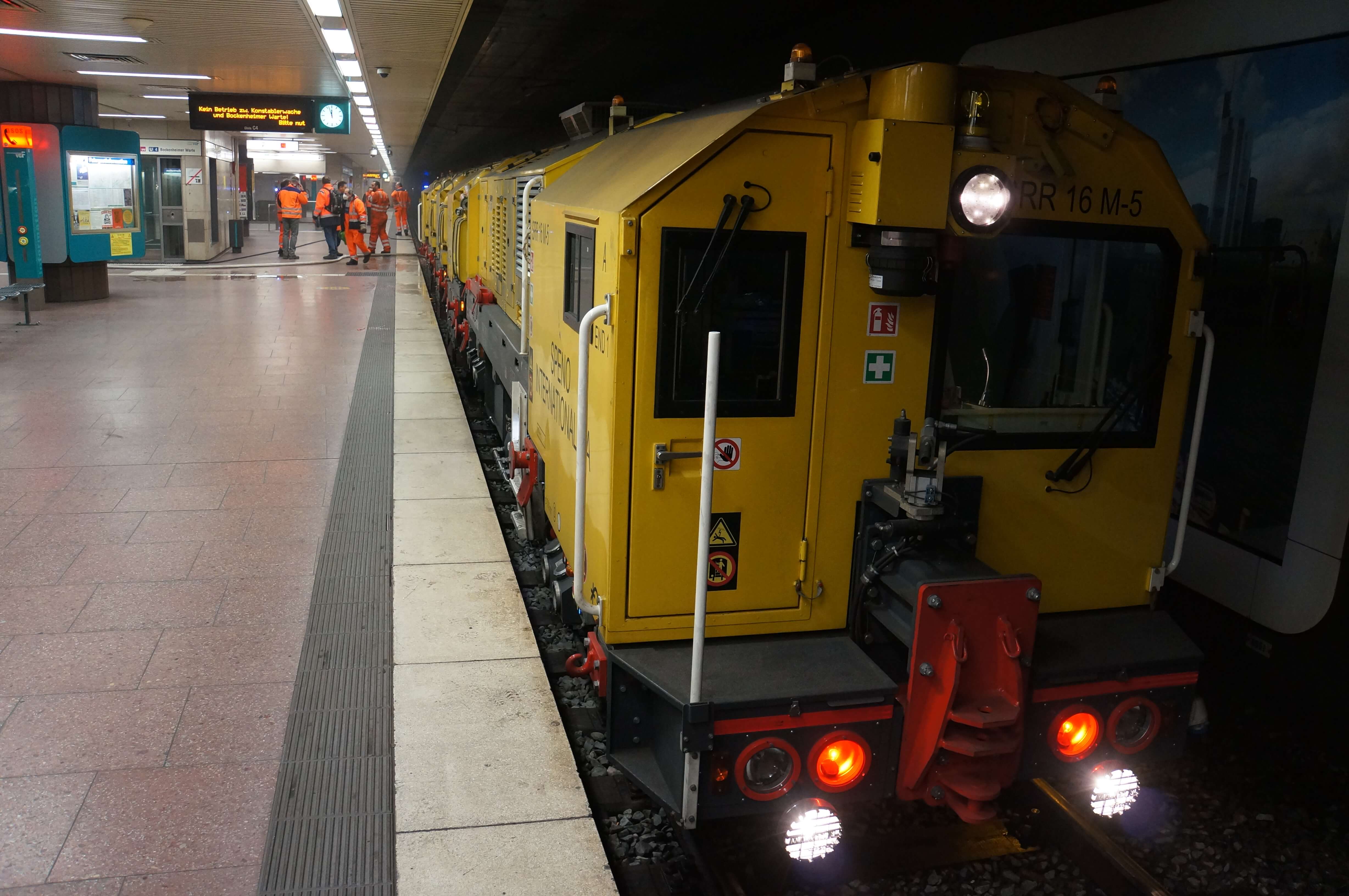 Schienenschleiffahrzeug an einer unterirdischen Haltestelle an der Personen in orangener Arbeitskleidung stehen.