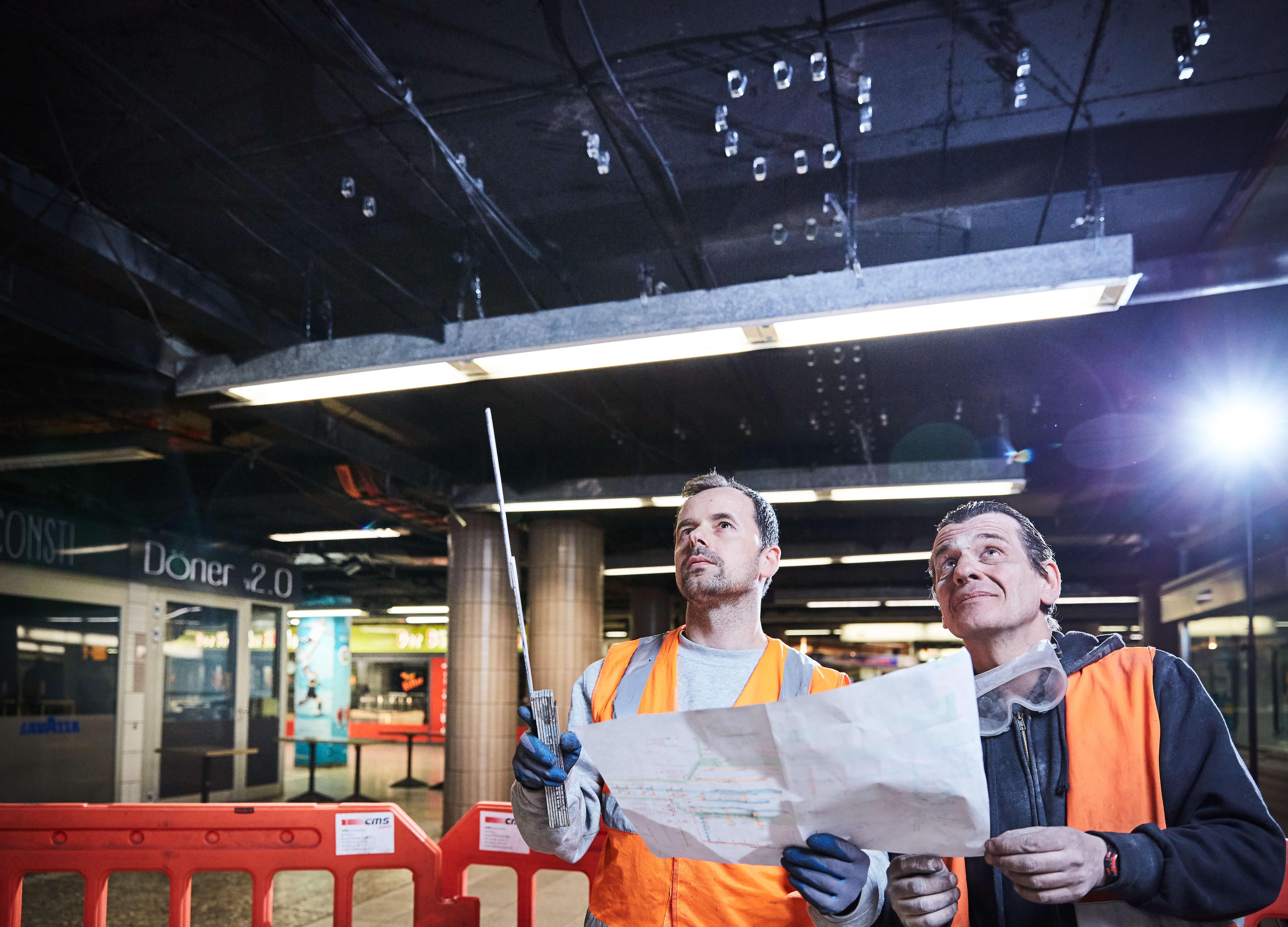 Zwei Männer in Warnwesten Metermaß und Plan in der Hand schauen an die Decke der B-Ebene