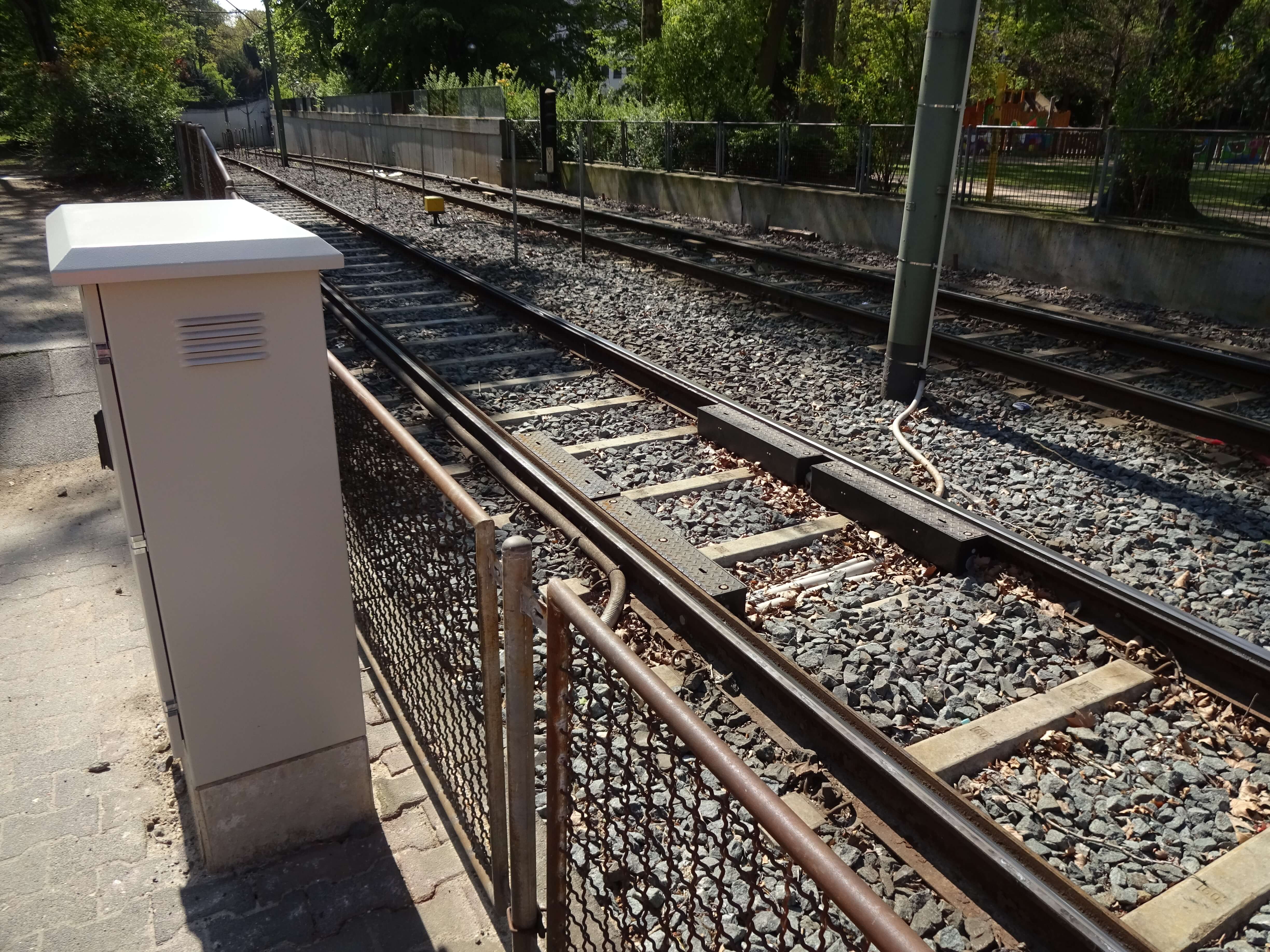 Schienen zwischen Zäunen und begrünter Umgebung und Stromkasten daneben.