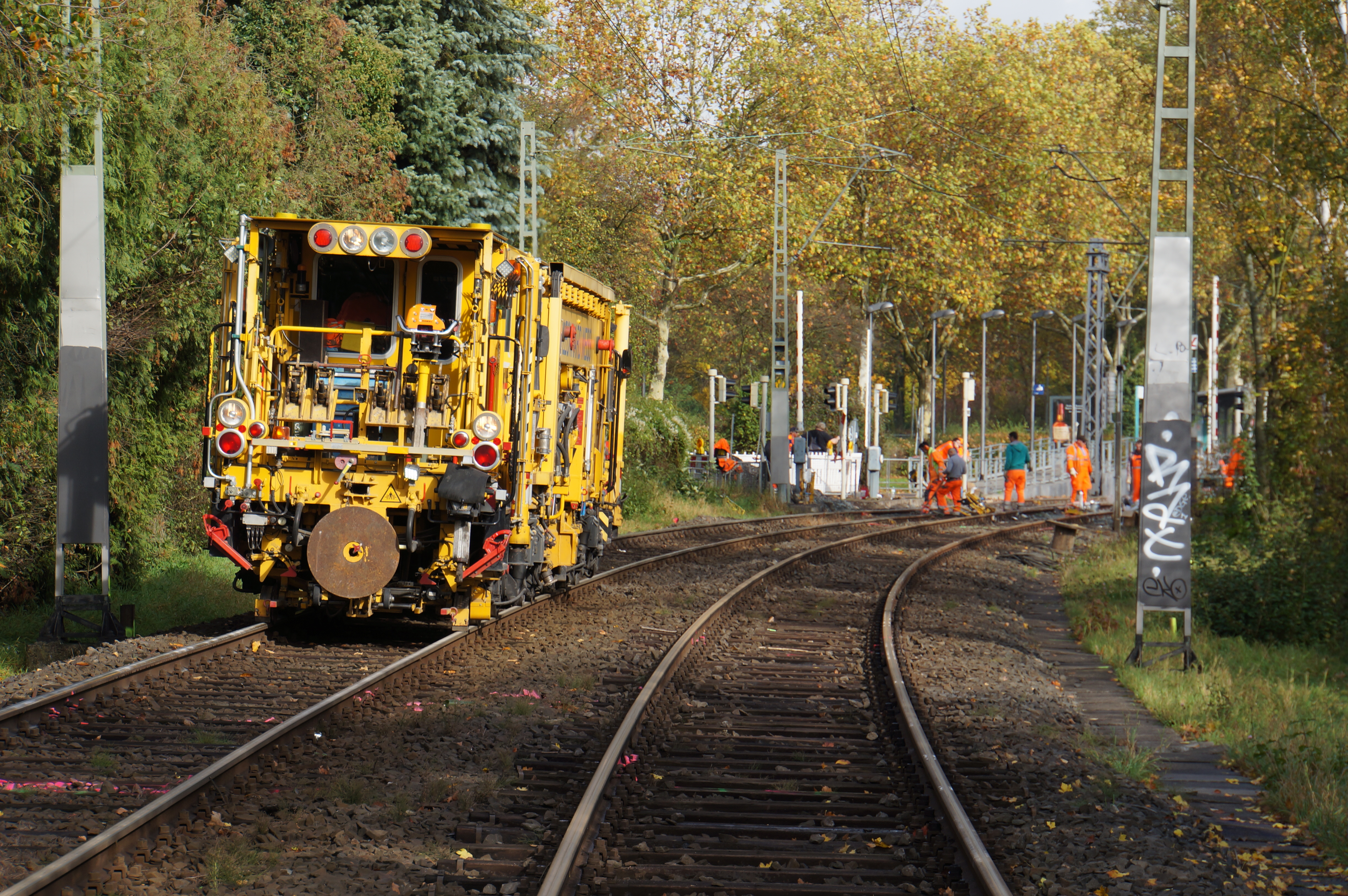 Gleisstopfer in grüner Umgebung, weiter hinten Bauarbeiter auf den Schienen