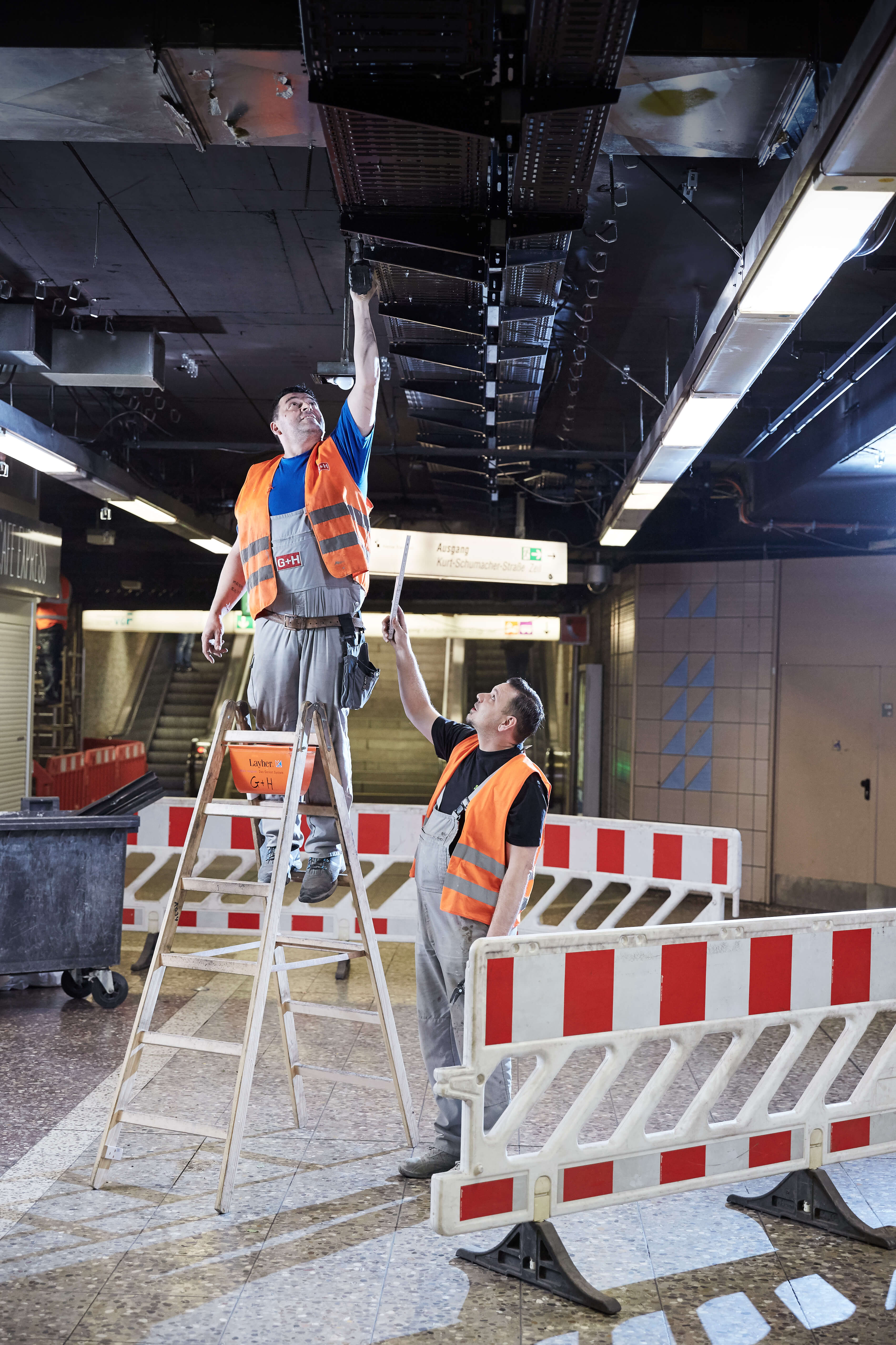 Zwei Männer in Warnwesten, der eine auf einer Leiter mit Akkuschrauber in der Hand, der andere daneben reicht Metermaß an und schaut zu.